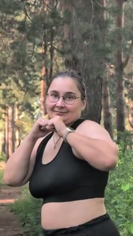 This is a short video clip of a young woman outdoors, seemingly in a wooded area. She is wearing a black sports bra and black pants. The woman has dark hair pulled back and is wearing glasses. She is in a boxing stance, with her fists raised. The background consists of tall trees and green foliage, suggesting a natural, possibly park-like setting. The lighting indicates it is daytime, likely late afternoon or early evening due to the soft light and shadows. The woman appears to be smiling or laughing, and her body language suggests she is actively engaged in a physical activity, possibly practicing martial arts or simply playing. There are no visible text elements in the video. The overall mood is lighthearted and energetic.