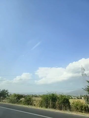 The video is a roadside view under a clear blue sky with scattered white clouds. A road is visible in the foreground, with its edge marked by white lines. To the right of the road, there is a grassy embankment with green bushes and some trees. In the distance, there is a dry, hilly landscape with some sparse vegetation and a few buildings. The sky is a prominent feature, with a faint contrail visible. No people or specific actions are discernible.