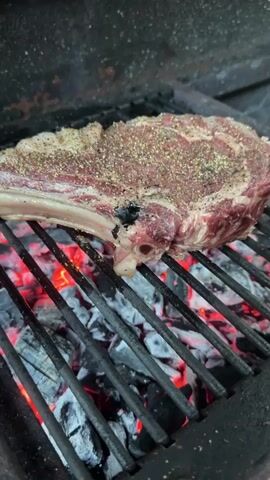 A raw steak seasoned with pepper sits on a grill over glowing red coals. The camera pans slightly to the right, showing the steak sizzling on the grill grates. The steak is thick and appears to be a ribeye cut. The grill is made of black metal. The coals are bright red and emitting heat. The background is dark and out of focus.