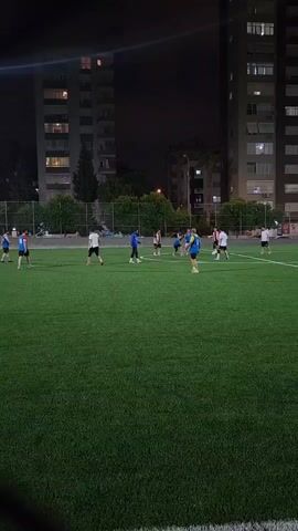 A group of people are playing soccer on a green artificial turf field at night. There are tall apartment buildings in the background, illuminated by lights. The players are wearing various colored shirts and shorts, and some are actively kicking and chasing a soccer ball. The scene appears to be a casual game or practice session.