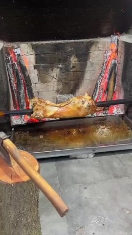 A whole lamb is being roasted on a spit over a fire in a brick fireplace. The fire is burning brightly on either side of the lamb, and the lamb is slowly turning. A metal tray underneath catches drippings. An axe rests on a wooden stump in the foreground. The video is taken from a stationary position, focusing on the roasting process.