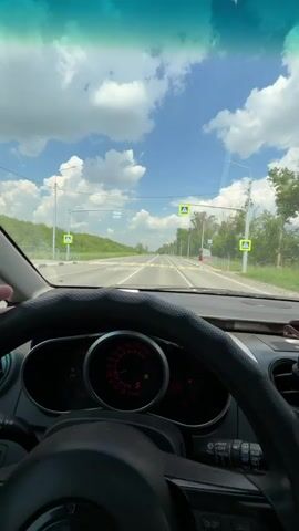 The video is filmed from the perspective of someone driving a car. The driver's hands are visible in the foreground, making a peace sign with their right hand and a "rock on" gesture with their left hand. The steering wheel is also visible, and the dashboard shows a speedometer with the needle indicating a speed between 0 and 5. The road ahead is a two-lane highway with a pedestrian crossing sign visible in the distance. The sky is blue with fluffy white clouds, and trees line the sides of the road. The sun appears to be shining brightly. The video appears to be taken during the daytime.