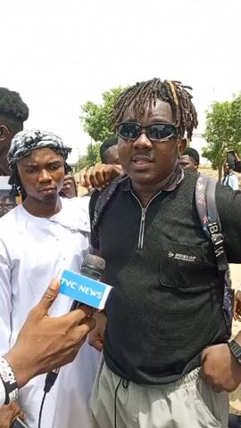Here is a concise description of the video:

A man with dreadlocks and sunglasses is being interviewed by a reporter holding a microphone with the TVC NEWS logo. The man is wearing a black shirt with a zipper and a backpack. He is surrounded by other people, some of whom are holding a white banner with black lettering. The environment appears to be outdoors, with trees visible in the background.