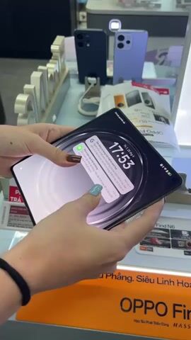 The video shows a person holding a black folding phone in their hands. They are folding and unfolding the phone. The phone displays a clock and some text in Vietnamese. Behind the person, there are other phones and promotional materials for Oppo phones. The environment appears to be a store display.