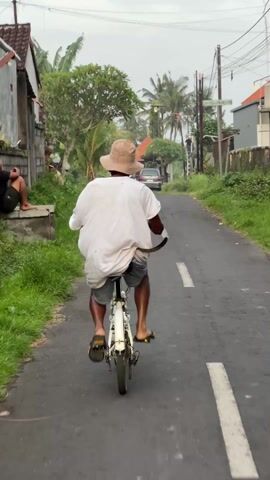 The video shows a man riding a white bicycle down a paved road. The man is wearing a light-colored bucket hat, a white T-shirt, and denim shorts. He is also wearing dark-colored sandals. The road has a white line down the center and green grass on either side. There are buildings, palm trees, and power lines in the background. The sky is overcast. The camera follows the man from behind as he pedals the bicycle.