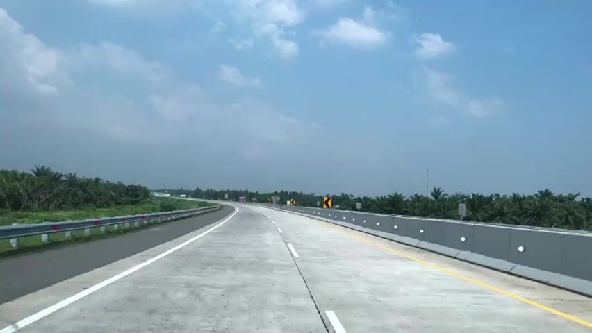 The video shows a highway with multiple lanes and a clear blue sky with scattered clouds. On the left side of the highway, there is a metal guardrail and a row of trees. On the right side, there is a concrete barrier with white circular reflectors and a speed limit sign indicating 40. The road appears to be empty except for a few vehicles in the distance. The landscape on both sides of the highway is characterized by palm trees, suggesting a tropical or subtropical region. The video is filmed from the perspective of someone driving on the highway, looking forward.