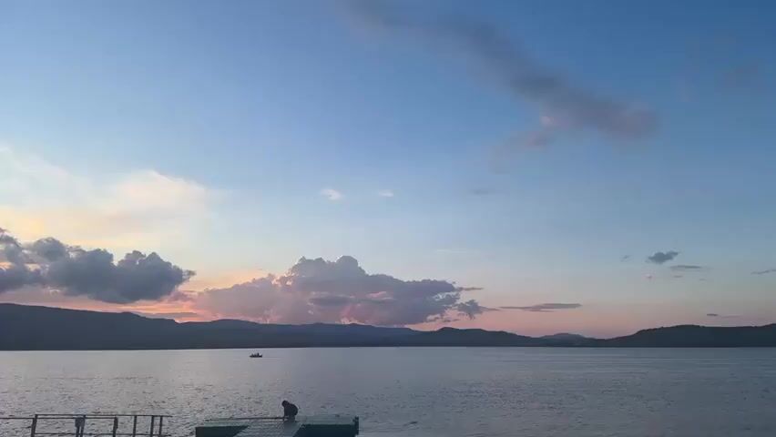 The video shows a wide shot of a calm body of water, likely a lake or sea, at dusk. The sky is a blend of blue, pink, and orange hues, with scattered clouds. In the foreground, a person is sitting on a floating dock. In the mid-ground, a small boat is visible on the water. The horizon is lined with distant hills or mountains. The overall scene is serene and captures the beauty of a sunset or sunrise over a natural landscape.