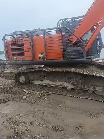 The video shows an orange excavator with a black cage on top, parked on a muddy, unpaved road. The excavator has tracks that are covered in mud. In the background, there is a body of water, possibly a lake or river, and some vegetation. The sky is overcast. The camera appears to be mounted on a vehicle or held by a person, and it pans across the scene. The road is rutted and appears to be under construction or repair.