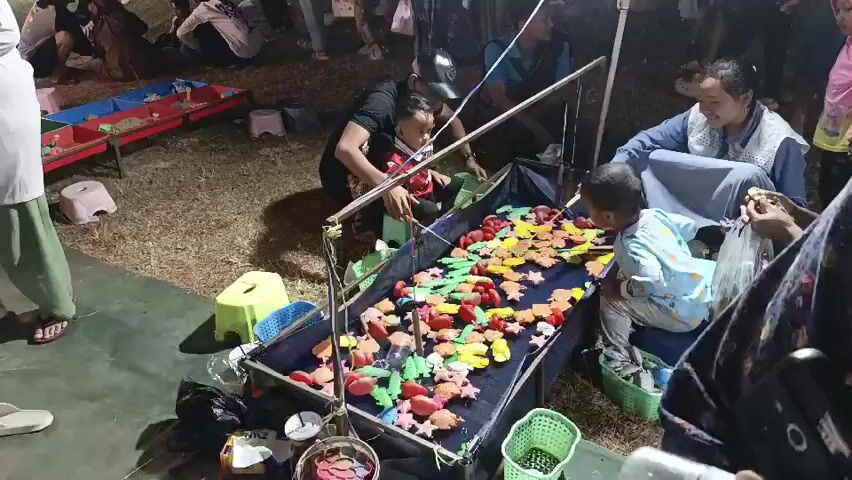 The video shows a group of people gathered around a fishing game stall at night. The stall is a rectangular container filled with water and numerous colorful plastic toys, predominantly sea creatures like fish, octopuses, and turtles. The toys are floating on the surface. A young boy, wearing a blue shirt with a dinosaur pattern, is sitting on a stool, actively participating in the game. He holds a fishing rod with a line and a hook, attempting to catch the floating toys. To his right, an adult woman, wearing a light blue vest over a white top and a headscarf, is seated and appears to be supervising or assisting the boy. She is also holding a fishing rod and observing the game. Next to her, another young child in a red shirt is also present and seems to be interacting with the game. In the background, other individuals are visible, some watching, others walking around the fairground. The ground is covered in dry grass or dirt, and there are some scattered plastic stools and buckets around the stall. A bright light is illuminating the game area, suggesting it is an outdoor night market or fair. The overall atmosphere appears festive and engaging.