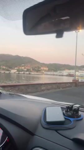 The video shows a view from inside a car. In the foreground, the dashboard and steering wheel are visible, along with a hand on the steering wheel. A phone is mounted on the dashboard. In the background, a body of water is visible, with buildings and hills on the other side. The sky is light pink, suggesting it is either sunrise or sunset. The car appears to be moving slowly.