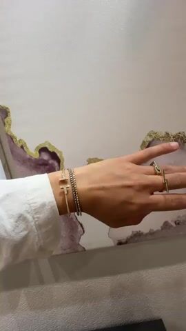 A person's left hand and arm are shown in profile, resting against a wall with abstract artwork. The person is wearing a white long-sleeved shirt. On the wrist, there are two bracelets. The first bracelet, closer to the hand, is a thin gold cuff with two small 'T' shapes, each adorned with tiny stones. The second bracelet, stacked above the first, is a beaded bracelet made of small, alternating silver and gold beads. On the fingers, two rings are visible. One is a thin gold band on the ring finger, and the other is a similar thin gold band on the index finger, forming an open geometric shape. The artwork behind the hand features streaks of purple and gold on a white background.
