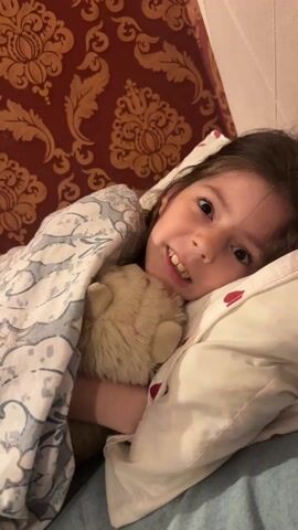 A young girl is lying in bed, nestled under a blanket with a pillow behind her head. She is hugging a beige-colored stuffed teddy bear. Her brown hair is slightly messy. The background is a red and gold patterned wall. The girl is saying “kiss, kiss, kiss,” as she holds the teddy bear close to the camera. She then says, “nom, nom, nom.”
