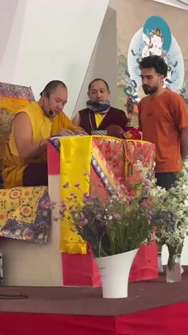 The video shows three people on a stage. Two are Buddhist monks wearing traditional robes and the third is a man with a beard wearing an orange t-shirt. One of the monks is sitting at a table covered in a colorful cloth and writing in a notebook. The other two are standing behind the table. There is a microphone attached to the first monk's ear. In front of the table are two vases of flowers. Behind them is a banner featuring an image of a deity. The stage is covered in a red cloth. The walls are white.