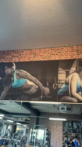 The video shows a mural in a gym. The mural is a black and white image of two people, a woman and a man, both wearing blue tops and working out with weights. The mural is mounted above a brick wall, with the rest of the gym visible below. There are various exercise machines and equipment in the gym, and at least one other person working out.