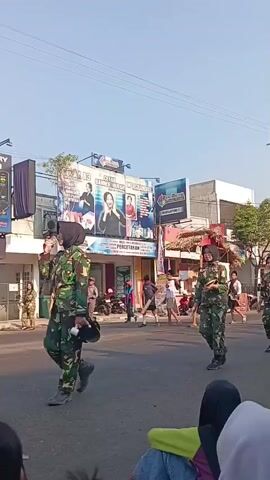 The video captures a street scene with several individuals in camouflage military uniforms marching. One uniformed individual in the foreground on the left is holding a rifle. Further down the street, another uniformed individual is visible. On the right side of the street, two more uniformed individuals are walking. A police officer in a high-visibility vest is standing in the middle ground, facing away from the camera. Behind the marching figures, there are buildings with billboards and other signs. A few onlookers are partially visible in the foreground, seated on the ground. The scene appears to be outdoors during the day, with a clear sky.