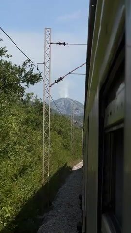 The video is a first-person perspective from inside a moving train. The camera is positioned to look out the right side of the train. In the foreground, the side of the train car is visible, along with a window. To the left of the train, railroad tracks curve away into the distance. Adjacent to the tracks is a steep embankment covered in dense green foliage. Several tall, metal utility poles with wires strung between them are visible along the embankment, indicating an electrified railway. In the background, a mountain range rises above the trees. At the summit of one of the mountains, a plume of white smoke or steam is rising into the clear blue sky. The sun appears to be shining, casting shadows and highlighting the scene. The train is clearly in motion, as evidenced by the blurred foliage and the perspective shift.