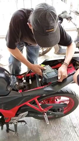 A person wearing a black t-shirt, blue jeans, and a black baseball cap is leaning over a black and red motorcycle. The person is wearing a black glove on their left hand and is holding a black electronic device connected to the motorcycle's undercarriage with their right hand. The motorcycle's seat is removed, revealing internal components. The person appears to be working on the motorcycle. The background is blurred but suggests an outdoor or garage setting with a concrete floor and some structures visible. The video is a close-up shot focused on the person and the motorcycle.
