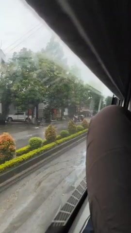 The video shows a view from a vehicle window looking out at a street scene. It is raining, and the road and trees are wet. There are several vehicles visible, including a white pickup truck, a red motorcycle, a tractor pulling a trailer, and two scooters. A person is riding the motorcycle, and two people are on one of the scooters. The street is lined with trees and a landscaped median with small, manicured bushes. Buildings and businesses are visible on the opposite side of the street. The camera appears to be moving, causing some motion blur.