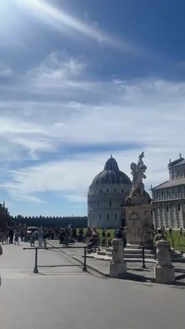 The video shows the Piazza dei Miracoli in Pisa, Italy. In the foreground, several people are visible, including one person wearing a hat and backpack, and another person wearing a red hat. A street vendor with a colorful array of items on their back is walking towards the camera. In the background, the Pisa Baptistery, the Cathedral of Pisa, and the Leaning Tower of Pisa are visible. The Cathedral of Pisa is partially covered in scaffolding. The sky is blue with some white clouds. The ground appears to be paved with stone. The video pans from left to right, revealing more of the square and its iconic structures.