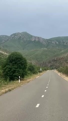 A point-of-view shot from a moving vehicle shows a two-lane asphalt road with a dashed white line in the center. The road curves slightly to the right in the distance. Lush green hills and mountains covered in trees and vegetation rise on both sides of the road under an overcast sky. A few small white roadside markers are visible.