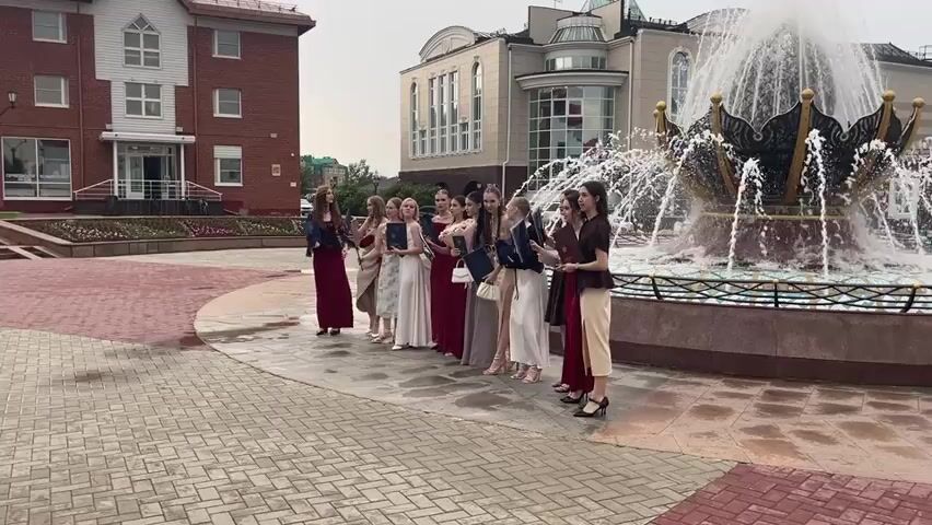 The video opens with a wide shot of a fountain with water spraying high into the air. A group of young women, dressed in formal attire and holding diplomas, are standing in a line in front of the fountain. The women are holding their diplomas in various ways, some are holding them in their hands, and others are holding them under their arms. The women are all looking towards the left side of the frame, where a group of people are standing and talking. The people on the left are dressed in casual clothing, and some of them are holding phones. The background of the scene is a brick building with a white trim and several windows. The ground is paved with cobblestones. As the video progresses, the camera pans to the left, revealing more of the group of people. A woman in a brown outfit is speaking to another woman in a light blue suit. A man in a black shirt is standing next to the woman in the blue suit, and he is holding a phone. The woman in the blue suit is holding a phone in her hand and appears to be looking at it. The camera then pans back to the right, where the young women are still standing in front of the fountain. The video continues to show the group of young women and the people on the left for a few more seconds before the video ends.