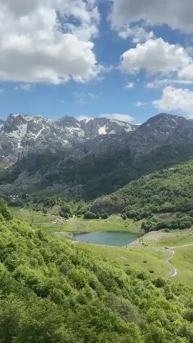 The video shows a picturesque mountain landscape under a partly cloudy sky. In the distance, a series of rugged mountains are visible, some with patches of snow on their peaks and slopes. The mid-ground is filled with lush green hills covered in trees and grassy meadows. Nestled among the hills is a small, serene lake with a tranquil blue-green surface. A winding road can be seen meandering down the hillside towards the lake. Scattered across the meadows and hills are a few small buildings, presumably part of a rural settlement. The overall impression is one of peace and natural beauty.