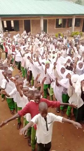 This is a video of a large group of children, seemingly in a school setting, performing a synchronized dance or activity. The children are mostly wearing white shirts and green pants, with some of the girls wearing hijabs. They are arranged in several lines, facing the camera, and are all raising their arms up and down in unison. The background shows a building that appears to be a school, with classrooms visible. The ground is dirt or gravel. The overall atmosphere appears to be energetic and joyful.