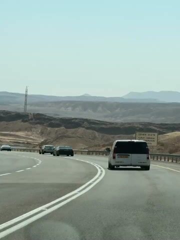 A white van drives on a highway with other cars. The van is in the right lane, and several cars are in the left lane. The highway is surrounded by desert landscape with mountains in the background. The sky is clear and blue. A sign on the right side of the road reads "DANGEROUS CURVES" in English.
