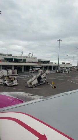 The video shows an airport tarmac from the perspective of inside an airplane, looking out the window. The window displays the airplane's wing with a pink and red stripe on it. Outside, several airport vehicles are visible, including white mobile stairs and a small white truck pulling two carts full of luggage. The airport terminal building is in the background, with glass windows and several people visible on a balcony. The sky is overcast, and the overall environment suggests a busy airport scene.