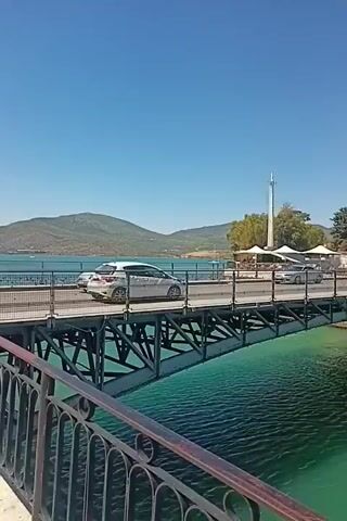 The video shows a body of water with a bridge crossing it. Cars are seen driving across the bridge. On one side of the water, there are buildings and trees, with a hillside in the background. On the other side, more buildings are visible along the coastline, as well as mountains in the distance. The water is a clear turquoise color, and the sky is a bright blue. The video appears to be taken on a sunny day. The camera pans across the scene, offering a panoramic view of the bridge, the water, and the surrounding landscape. A railing is visible in the foreground of some shots.