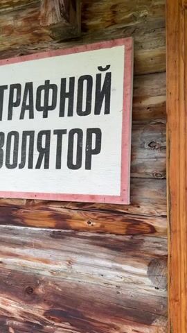 The video begins with a close-up of a weathered wooden wall with a partial sign that has Cyrillic text. The camera then pans to the right, revealing a narrow wooden hallway with several doors and an open wooden door on the right. The hallway is dimly lit, with wooden paneling on the walls and ceiling, and wooden floorboards. There are some metal doors visible down the hallway, and a taxidermied animal head mounted on the ceiling. As the camera continues to pan, it focuses on a room with wooden walls and a wooden floor. Several military-style jackets and pants are hanging on the wall. A small wooden table with a metal cup and a pair of black boots are in the foreground. The lighting in the room is warm and natural.