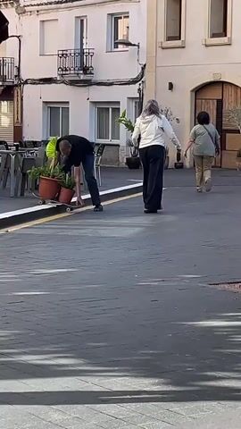 The video shows a man adjusting two flower pots on a skateboard in a public square. He is wearing a black t-shirt and dark pants. Two women are walking away from him in the background. An elderly person in a gray cardigan, jeans, and Crocs walks past the camera. The setting appears to be a European town with light-colored buildings, outdoor cafe seating, and cobblestone paving.