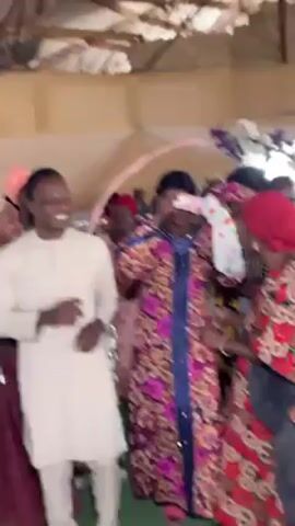 The video captures a joyous moment at what appears to be a wedding reception or similar celebration, taking place indoors. A group of people are gathered, predominantly women dressed in vibrant, patterned dresses and head coverings, indicating a cultural event. At least one woman is wearing a blindfold and holding a gift bag, suggesting a game or tradition.  A man in a white outfit stands among the crowd, smiling and interacting with the blindfolded woman. The environment includes a decorated archway and general festive setup, suggesting an atmosphere of celebration and community.