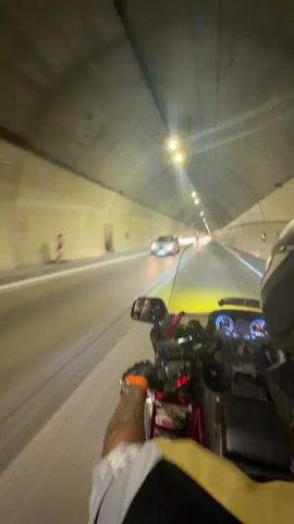 The video is a first-person view from a motorcycle traveling through a tunnel. The rider is wearing a helmet and a black and yellow jacket. They are holding the handlebars with gloved hands. On the motorcycle, there is a red and chrome fairing, a windshield, and various gauges and controls visible. Other vehicles, including cars, are also visible in the tunnel, moving in the same direction. The tunnel has a concrete ceiling with lights spaced at intervals, and the walls are also made of concrete. The lighting in the tunnel creates streaks of light due to the motion. The road appears to be paved and has lane markings.