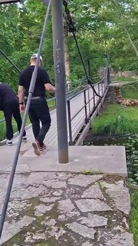 A group of four people are walking across a suspension bridge. The bridge is made of wood and metal and is surrounded by trees and foliage. The people are walking from left to right in the frame. The person on the far left is wearing a dark jacket and jeans. The person next to them is wearing a black tracksuit. The third person is wearing a black t-shirt and black pants. The fourth person is wearing a black jacket and black pants. The bridge appears to be in a park or natural setting. The time of day is daytime.