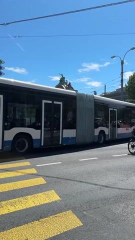 A bus with multiple doors and sections is driving on a street in front of a crosswalk. There is a building with a statue on top in the background, as well as trees and utility poles. The sky is blue with some clouds.