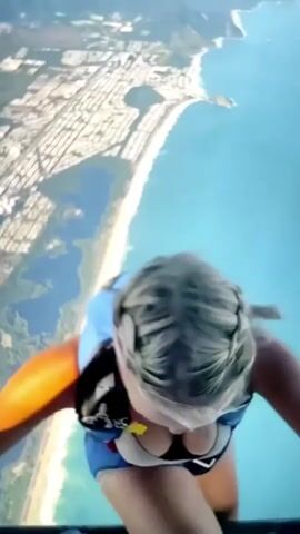 A first-person perspective video shows a person with blonde hair in a blue and black jumpsuit with a parachute strapped to their back. They are leaning forward over a ledge, with their hands gripping the edge. Below them is a coastline with a beach and buildings, extending out to a bright blue ocean. The camera is angled downwards, giving a dizzying view of the drop. The video is shaky, suggesting movement and perhaps excitement or nervousness. Text overlays at the bottom of the screen include hashtags like #skydiving, #jumping, and #viral, as well as what appears to be a username or handle.