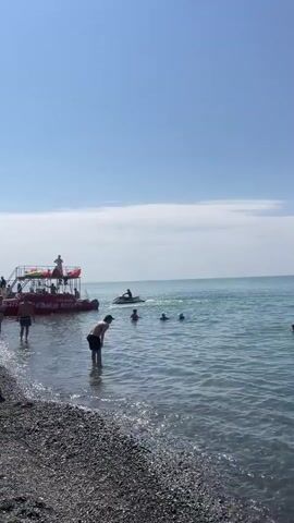 The video shows a crowded beach scene on a sunny day. The beach is covered in pebbles, and the sea is calm with gentle waves. Several people are in the water, swimming or playing near the shore. In the background, a red boat with multiple levels and people on it is docked or moving slowly near the beach. A person is operating a jet ski in the water, creating a wake. Children are visible on the beach, some playing near the water's edge. The sky is clear blue with a few scattered clouds.