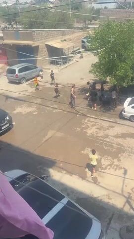 The video captures a scene on a street where several people, primarily children, are engaged in what appears to be a water fight. The ground is wet, indicating the use of water. There are cars parked on the street and in the background, as well as buildings and trees. The individuals are interacting with each other, with some holding buckets or containers that might be used for throwing water. One person is standing with their arms outstretched, and others are in various stages of movement, suggesting active participation in the water fight. The overall atmosphere seems playful and energetic.