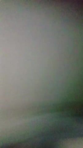 A young male with brown, messy hair and wearing a black t-shirt is seen in a dimly lit room. He is looking at the camera and making facial expressions. His right arm is visible, and he appears to be speaking or making sounds. The background is out of focus and appears to be a wall or ceiling.