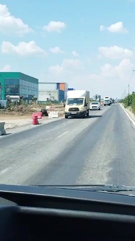 The video shows a view from inside a vehicle traveling on a road. Construction is evident on the left side of the road, with orange cones and barriers present. Several vehicles are seen on the road, including a yellow van, a white car, and various trucks. The sky is blue with scattered clouds, and there is greenery along the right side of the road. The footage appears to be captured during daylight.