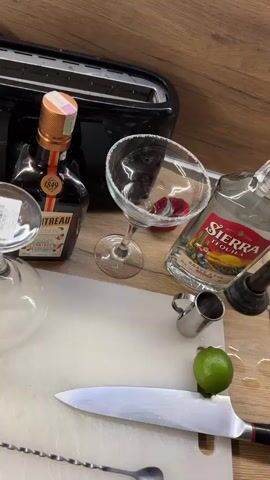 A person prepares a cocktail. They pour the contents of a metal cocktail shaker into a sugar-rimmed glass. Bottles of Cointreau and Sierra Tequila sit nearby, along with a lime and a knife on a white cutting board. A toaster is visible in the background. The countertop is wood.