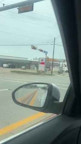 The video shows a street view from inside a car. The car is stopped at a traffic light in front of a 76 gas station. The gas price sign reads $3.99.  The street sign indicates "Cooper St". The car’s side mirror shows cars behind it on a street with yellow lines. The weather appears to be overcast. The person inside the car says "This the price of gas bruh!" in a surprised tone.