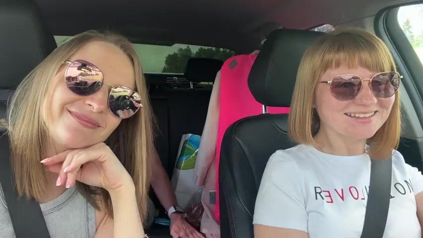 Three women are in a car. Two are in the front seats, and one is in the back. All three women are wearing sunglasses and seatbelts. The woman in the driver's seat has blonde hair. The woman in the passenger seat has short blonde hair with bangs. The woman in the back has long blonde hair. There are bags and clothing in the back seat behind her. The women are smiling and talking. The music is playing.
