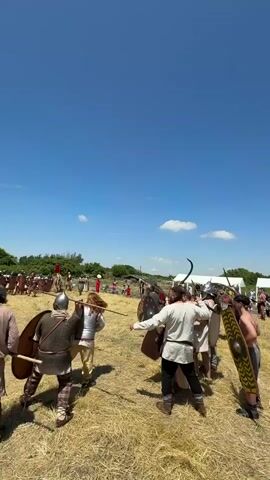 A group of people dressed in historical warrior costumes stands on a dry, grassy field under a bright blue sky with a few white clouds. In the foreground, three individuals face away from the camera, holding shields and weapons. One wears chainmail and a helmet, another a white tunic and trousers, and the third a light-colored tunic and trousers, holding a spear. To their right, several other warriors with helmets and shields are clustered together. In the midground, more costumed individuals are visible across the field, some in red and white attire, others with various weapons and armor. In the background, spectators are seated on the ground, and a white tent is partially visible on the right. The overall scene suggests a historical reenactment or festival.