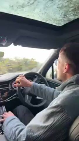The video shows the interior of a car, likely an SUV, with two people visible inside. The driver is a man wearing a grey patterned shirt. He is holding the steering wheel with both hands and appears to be navigating a turn on a country road. A woman with blonde hair sits in the passenger seat. The dashboard is modern with multiple screens displaying information like navigation. The car has a sunroof, which is visible. Outside, the scenery consists of green fields and trees under a sky that looks overcast or rainy. The video seems to be shot from the perspective of a camera held within the car, looking through the windshield.
