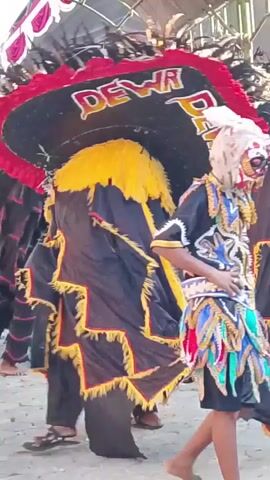 The video shows a close-up of a person wearing a large, elaborate costume. The costume appears to be a mask or headdress, black with a wide red border adorned with red and white trim. The word "DEWI" is written in white with a red outline on the front of the headdress, repeated twice. A bright yellow fringe hangs down from the front of the headdress. Black fabric with yellow zigzag trim drapes over the wearer's body, extending outward as they move. The person's legs are visible below the fabric, and they are wearing dark pants and sandals. In the background, other people are visible, some also wearing colorful costumes, suggesting a performance or parade. The environment appears to be outdoors, possibly under a canopy or tent structure. The lighting is somewhat diffused, and the camera is focused on the central figure, creating a shallow depth of field. The overall impression is one of movement and cultural performance.