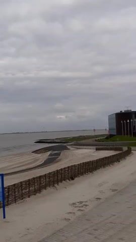 The video shows a beach on a cloudy day. The beach is mostly sand, with a woven fence running along the edge of the beach.  There is a body of water in the background, possibly an ocean or a large lake. A building is visible to the right of the frame. There are several vehicles, including a red truck and a trailer, parked on the beach. In the middle of the video, the clouds open up to reveal blue sky. There is a street light with a sign.