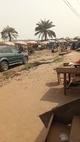 The camera is positioned in a market area, possibly a stall or storefront. The foreground includes wooden steps and a counter laden with goods, including red produce resembling peppers. A table in front of the steps also displays goods. The area in front of the stalls is a sandy or dirt road with visible trash. Across the road, a bustling market scene unfolds, marked by numerous individuals congregating under colorful umbrellas and stalls. A vehicle is parked at the edge of the road. Palm trees are scattered throughout the area, contributing to the warm, sunny environment. Power lines stretch overhead. The overall atmosphere is one of a busy, typical market day.
