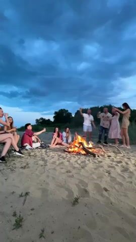 A group of eight people, including five women and three men, are gathered around a bonfire on a sandy beach under a dramatic, cloudy twilight sky. Three women are sitting on the sand, with two men standing behind the bonfire, and another three people positioned to the left of the frame. A man in a white shirt and light blue shorts raises his glass in a toast. Others raise their glasses as well. The fire is burning brightly, and there are trees and vegetation in the background.